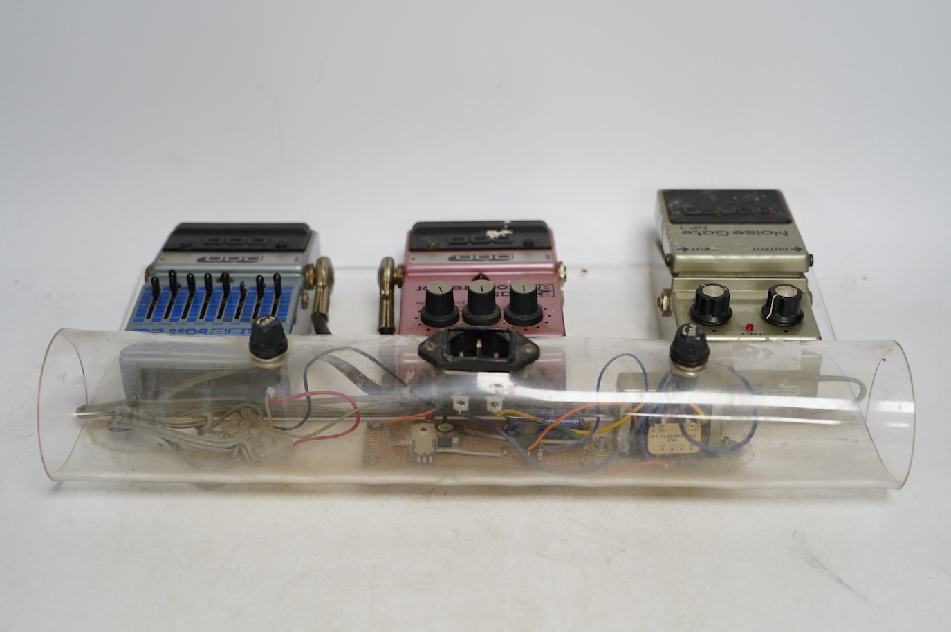 A bass guitar pedal board including; a DOD FX42 B bass EQ pedal, a DOD FX82 bass compressor and a Boss noise gate NF-1 pedal, mounted on a clear Perspex frame. Condition fair, some wear from use evident.